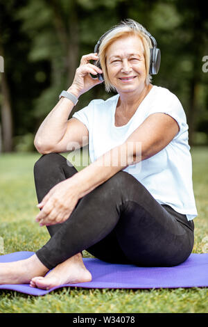 Happy senior femme à écouter la musique avec un casque tout en pratiquant le yoga dans le parc. Concept d'une vie active à la retraite Banque D'Images