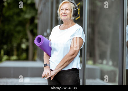 Portrait d'une femme senior actif dans des vêtements de sport avec tapis de yoga debout près du bâtiment à l'extérieur. Concept d'un mode de vie sain sur la retraite Banque D'Images