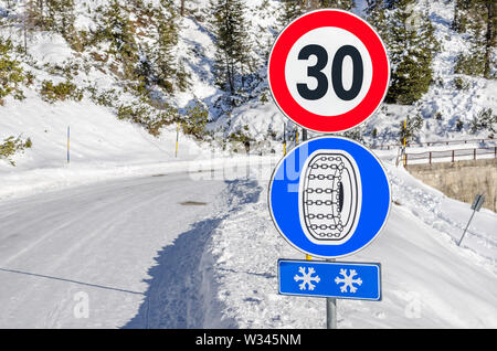 Chaînes à neige route obligatoire signer ci-dessous un signe de limite de vitesse le long d'une route sinueuse de montagne enneigée Banque D'Images