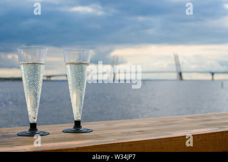 Deux verres de champagne en plastique sur une surface en bois sur l'arrière-plan de la ville, le grand pont, la mer et le ciel sombre. Banque D'Images