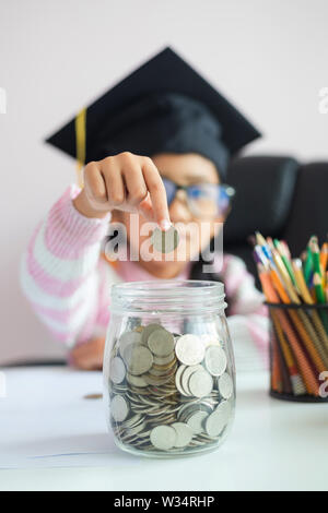 Peu d'Asian girl wearing hat diplômés mettre la pièce dans le bocal en verre clair tirelire et sourire avec bonheur pour de l'argent d'économie pour wealthness succes Banque D'Images