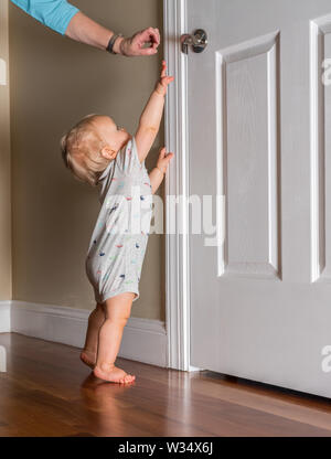 Jeune bébé juste capable de marcher jusqu'à la poignée de porte sur plancher en bois Banque D'Images