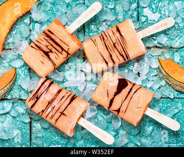 Vue de dessus des sucettes glacées faites de melon cantaloup avec du chocolat et des cubes de glace sur fond turquoise rustique. Banque D'Images