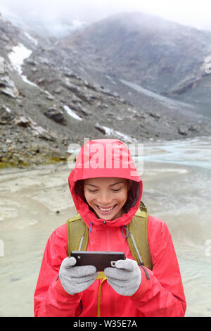 Téléphone intelligent woman texting on smartphone app sms en utilisant l'écran tactile avec des gants. Happy hiker with mobile phone en extérieur dans la nature dans la pluie. Fille avec du tissu conducteur de gants pour écran tactile. Banque D'Images