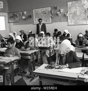 Années 1960, historique, un enseignant masculin avec des écoliers arabes assis sur des bureaux en bois dans une salle de classe, Arabie Saoudite. La plupart des jeunes garçons portent le traditionnel keffiyeh, foulards du moyen-Orient. Banque D'Images