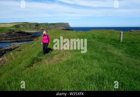 Woman Walking in Dunseverick Harbour avec Benbane Head en arrière-plan sur le Giant's Causeway chemin côtier, comté d'Antrim, en Irlande du Nord, Royaume-Uni Banque D'Images