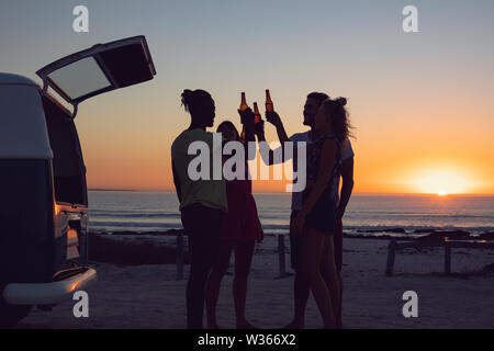 Friends toasting beer bottles près de camping-van pendant le coucher du soleil Banque D'Images