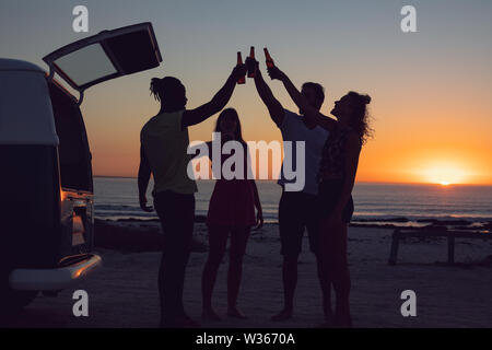 Friends toasting beer bottles près de camping-van pendant le coucher du soleil Banque D'Images