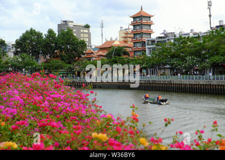 Deux travailleurs de l'assainissement s'asseoir sur le bateau, se déplacer sur canal le jour, paysage de bougainvilliers fleurissent en premier plan et arrière-plan, la pagode à Ho Chi Minh city Banque D'Images