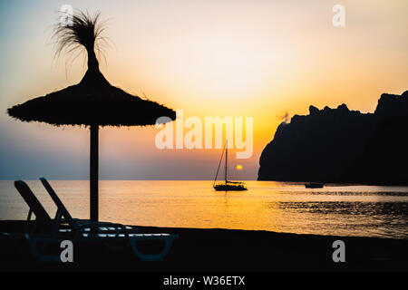 La silhouette du soleil de chaume avec des chaises longues à l'avant-plan de la silhouette d'un voilier au lever du soleil sur la Méditerranée Banque D'Images