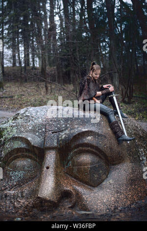 Fille avec une épée sur une tête en pierre dans la forêt sous la pluie Banque D'Images