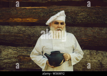 Chef cuisinier homme barbu en uniforme et chapeau à longue barbe sur le visage sérieux maintenant vieux fer à repasser plateau électrique journée ensoleillée en plein air, sur fond de bois Banque D'Images