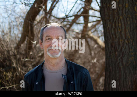 Homme portrait of mature man avec les cheveux gris et la barbe en veste de cuir sombre à l'extérieur debout près de l'arbre sur fond de ciel au début du printemps. Banque D'Images