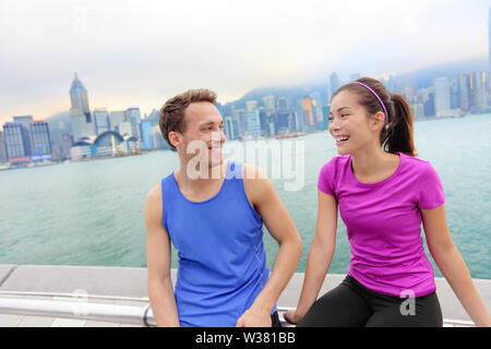 Porteur se détendre après entraînement à Hong Kong City. L'exécution du Caucase et de l'homme et la femme asiatique courir après le fait de prendre une pause de parler ensemble sur l'avenue des étoiles dans le port de Victoria, Hong Kong Skyline. Banque D'Images