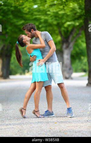 Premier baiser - jeune couple d'amoureux dans l'amour passionnément embrasser debout sur chemin dans parc d'été. Portrait complet du corps de l'homme de race blanche et asiatique femme en bleu robe d'aimer et d'étreindre l'autre. Banque D'Images