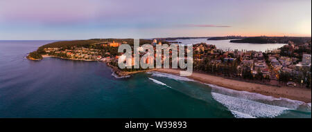 Les vagues de l'océan Pacifique roulant sur le sable de plage de Manly au coucher du soleil face à bord de l'eau et le port de Sydney et de la ville lointaine de la CDB. Banque D'Images