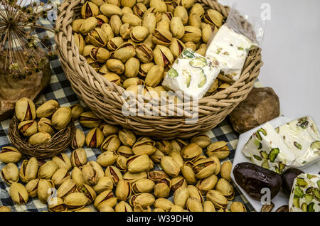Pistaches frit avec le kaki et le nougat dans un panier en osier et sur une assiette blanche à côté des pierres et des fleurs séchées sur papier avec carré bleu Banque D'Images