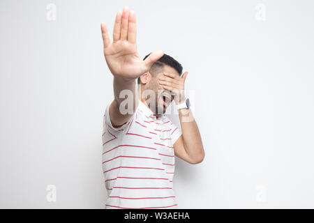 Stop, je ne veux pas examiner cette question. Portrait de jeune homme barbu confus en t-shirt à rayures, permanent couvrant ses yeux et d'arrêter en montrant le geste. Banque D'Images