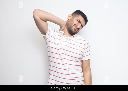 Douleur au cou ou la douleur. Portrait de beau jeune homme barbu en t-shirt à rayures debout et tenant son cou douloureux. Piscine studio shot, isolé sur blanc Banque D'Images