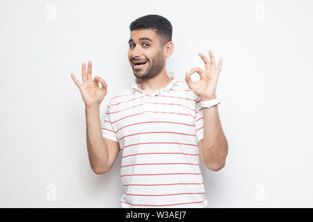 Portrait de funny beau jeune homme barbu en t-shirt à rayures avec permanent Ok s'identifier et à la recherche à l'appareil photo avec bonheur. Piscine studio shot, isolat Banque D'Images