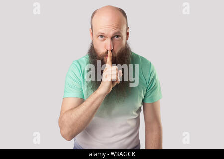 Être calme. Portrait d'âge moyen sérieux homme chauve avec longue barbe en vert clair t-shirt debout avec le doigt sur les lèvres et montrant le silence signe. indoo Banque D'Images