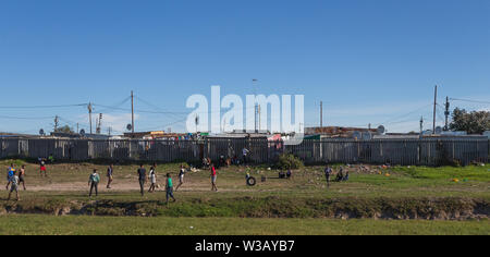 Groupe de pauvres enfants africains noirs lors d'une partie de soccer ou de football du côté de la route avec tin shack maisons dans l'arrière-plan Cape Town Banque D'Images