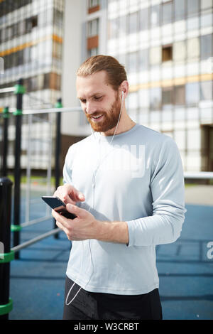 Heureux homme barbu beau chandail en comité permanent sur la formation et l'utilisation du sol pour les communications en ligne smartphone avec l'entraîneur Banque D'Images