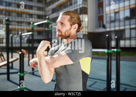 Jeune homme pensif grave avec barbe rouge debout sur l'équipement moderne, terrain de sport et d'étirement du bras de bicep Banque D'Images