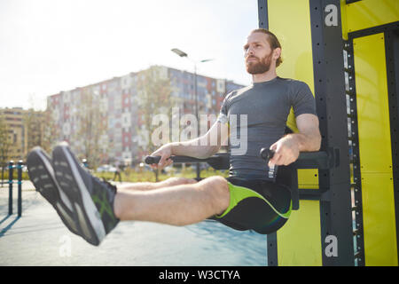 Jeune homme barbu concentré dans les vêtements de sport accroché sur les barres parallèles et d'exercer sur les bars en plein air Banque D'Images