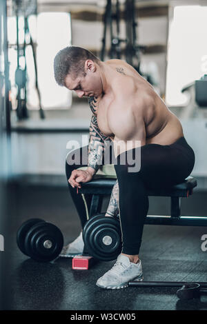 Bodybuilder motivés guy est assis sur un banc et effectue des exercices pour ses biceps. Banque D'Images