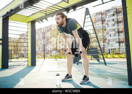 Assez jeune homme barbu avec barbe appuyée sur les genoux tout en regardant souffle après l'exercice intense sur le terrain de sport Banque D'Images