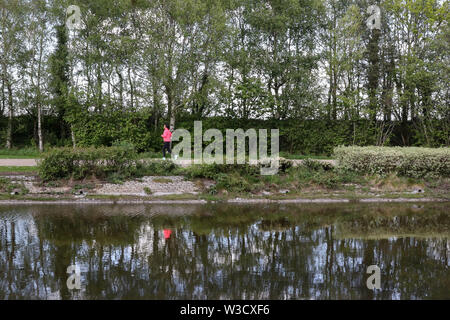 La réflexion d'une femme jogger rose en haut et guêtres noires tournant autour du lac du parc au parc Victoria, Belfast, Irlande du Nord. Banque D'Images