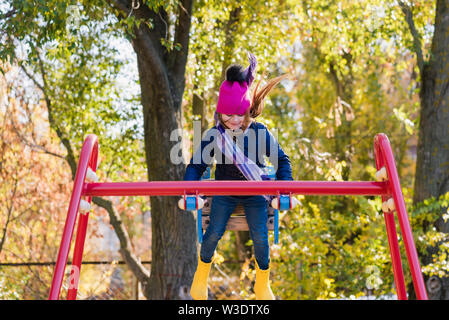 Girl in pink hat ride haut sur swing. Cute child à l'aire de jeux au cours de l'automne de marche Banque D'Images