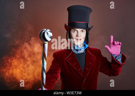 L'homme cirque fabuleux dans un chapeau et un costume rouge posant dans la fumée de couleur sur un fond sombre. Un clown lors d'une fête, l'homme représentant d'un conte de fées. Wiz Banque D'Images