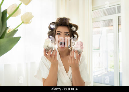 Belle jeune femme brune dans les bigoudis tenant deux cupcakes à deux mains debout dans cuisine lumineuse à la surpris Banque D'Images