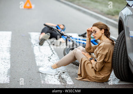 Femme triste assis près de la voiture, inquiets de l'homme blessé gisant sur le passage pour piétons après l'accident de la route Banque D'Images