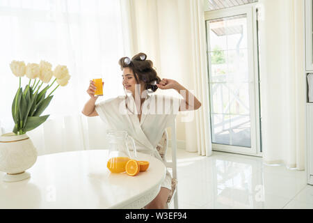 Jolie jeune femme brune dans les bigoudis bénéficiant d'orange jus fraîchement pressé qui s'étend après le sommeil en cuisine lumineuse Banque D'Images