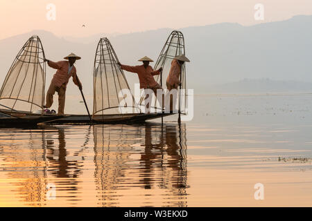 Inle, Myanmar - Mars 2019 : trois pêcheurs birmans traditionnels d'aviron de la jambe au lac Inle au lever du soleil, tenant dans les mains. filets Silhouette rétro-éclairage ref Banque D'Images