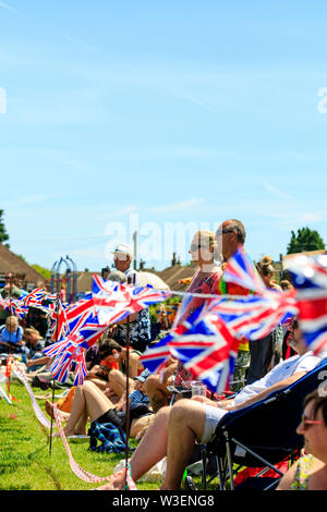 Union Jack noir voletant dans la brise avec rangée de gens debout et assis à regarder l'événement invisible. Un soleil brillant. L'espace négatif. Banque D'Images