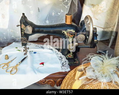 Une très vieille machine à coudre à pédale en noir avec décor, entouré par des ustensiles de couture, une nappe, un chapeau de paille avec ruban de tulle et rideaux délicats Banque D'Images