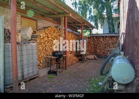 Un tas de bois au mur de la maison avec un grill et le réservoir d'eau. Le bois de chauffage est couverte d'un toit. Le fort et mensonge. Accrocher la jante des roues. Banque D'Images