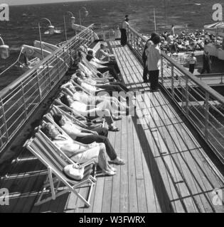 Années 1960, historique, dans l'océan, les passagers adultes sur le Sagafjord cruise ship assis, la détente au soleil sur des chaises longues à l'extérieur sur la terrasse en bois du navire. Banque D'Images