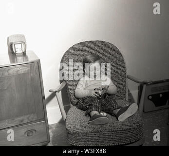 Années 1960, historiques, à l'intérieur d'une chambre, jeune garçon assis dans une chaise de salon, jouant avec une caméra et avec une radio sans fil de l'époque sur le sol à côté. Banque D'Images