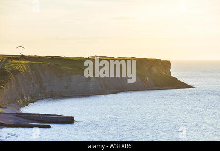 Falaises rocheuses couvertes d'herbe verte au coucher du soleil. Un homme pratiquant le parapente sur eux. Normandie, France. Banque D'Images