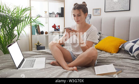 Happy young girl sitting on bed using smartphone et ordinateur portable à la maison. Les gens, la technologie, les loisirs, le magasinage en ligne concept Banque D'Images