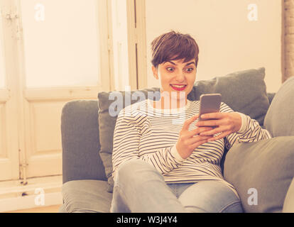 Happy young woman on smart mobile phone chat dans les applications de médias sociaux assis sur le canapé à la maison dans les gens et le mode de vie sociale, lance une Banque D'Images