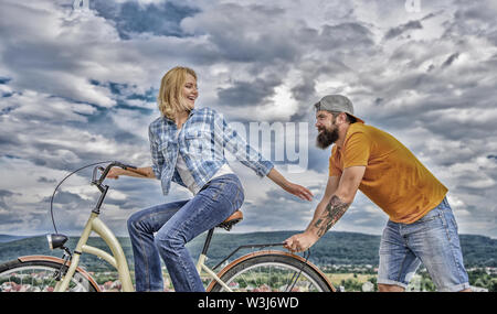 Homme pousse fille faire du vélo. Vélo fille tandis que l'homme la soutenir. Aide à croire en vous-même. Impulsion L'impression de commencer à bouger. Des promenades en vélo femme fond de ciel. Pousser et la promotion. Impulsion à se déplacer. Banque D'Images