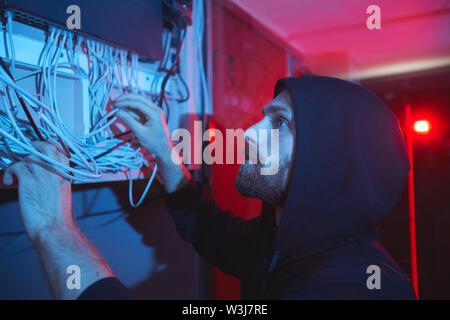 Concentré grave jeune spécialiste réseau barbu in hoodie debout dans une pièce sombre et d'examiner les câbles du server Banque D'Images