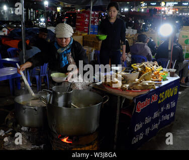 Diners assis sur des tables et des chaises en plastique, manger du poulet porridge au restaurant en plein air, marché alimentaire nocturne chaudron de nourriture sur poêle à charbon par temps froid Banque D'Images