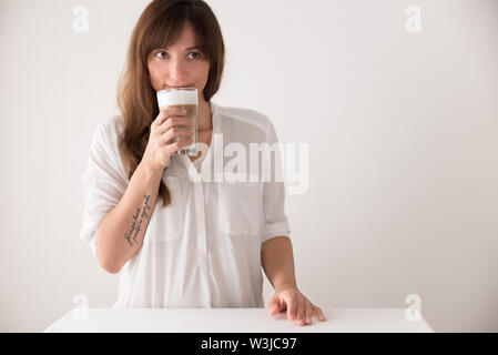 Jeune brunette woman contre fond blanc de boire du café latte macchiato et souriant Banque D'Images